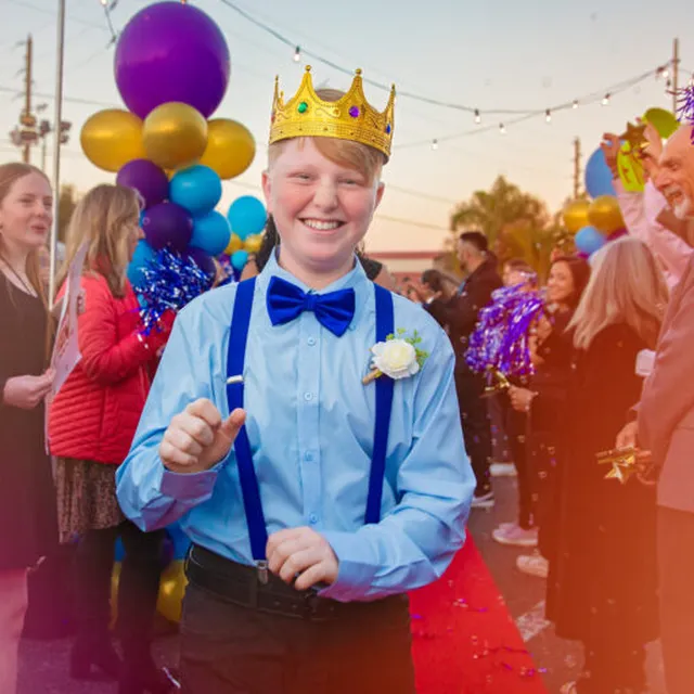 A smiling young man in a gold crown and blue bow tie walking a red carpet as friends cheer