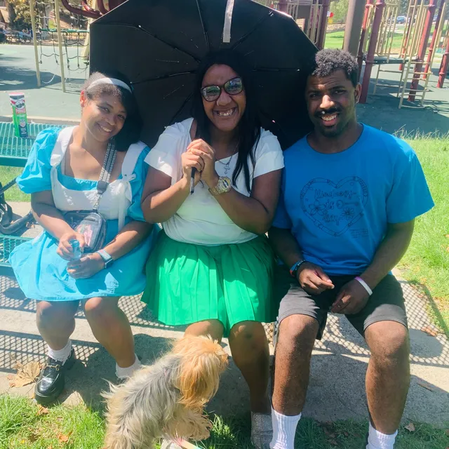 Three friends smiling on a park bench under an umbrella, with a little dog