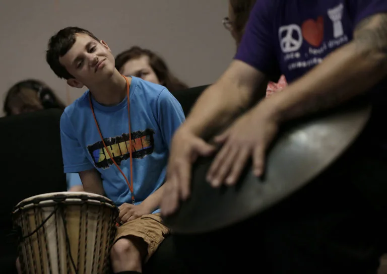 A young man smiling as he listens and plays along on a hand drum