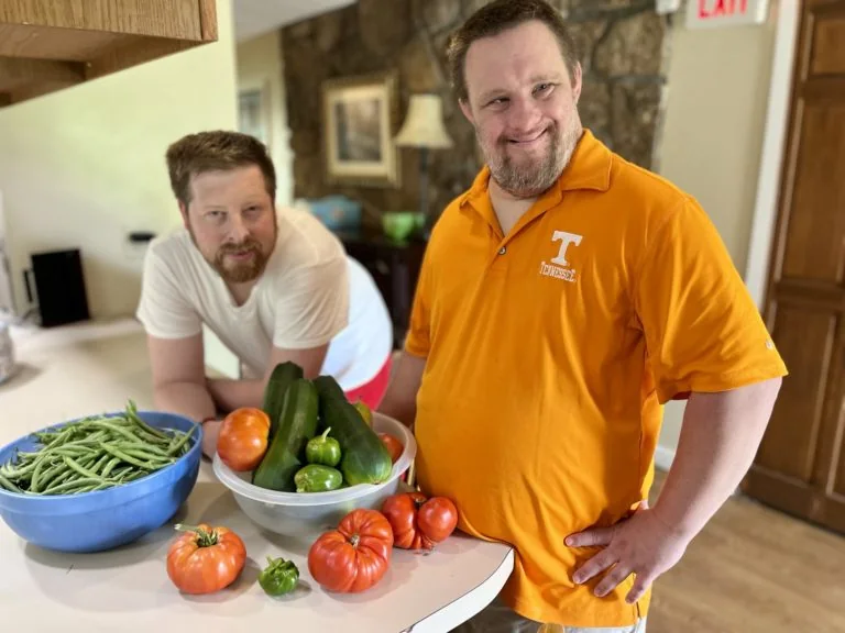 Two friends smiling beside a kitchen counter full of fresh garden vegetables