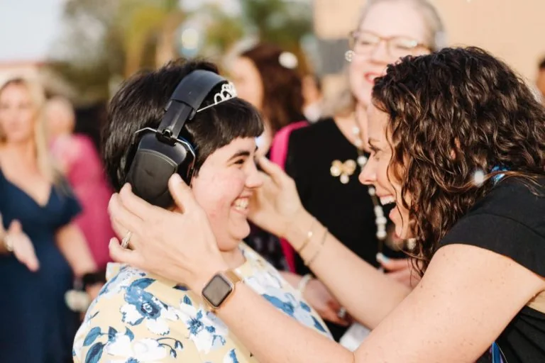 A smiling friend wearing headphones and a tiara as a friend leans in, laughing with joy