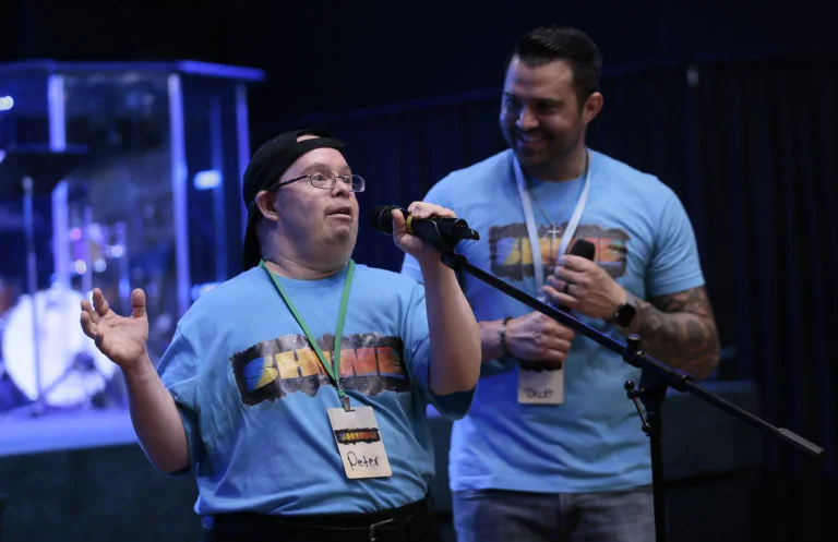 A friend speaking into a microphone on stage while a smiling friend stands beside him